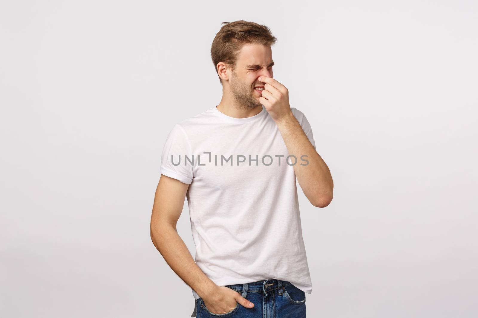 Disappointed and disgusted blond handsome caucasian man close nose with fingers, squinting and turn away from aversion and disgusted, smell something bad, product stinks, standing white background by Benzoix