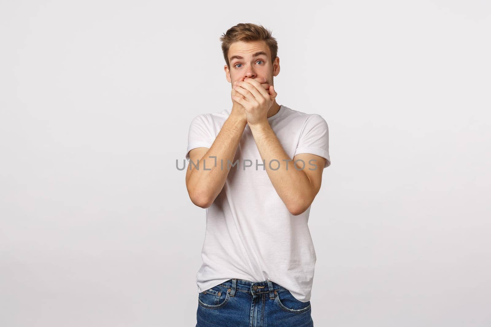 Shocked concerned young blond guy found out dirty secret, gossiping, shut mouth, press palm to lips and stare camera speechless, astounded witnessing something, white background.