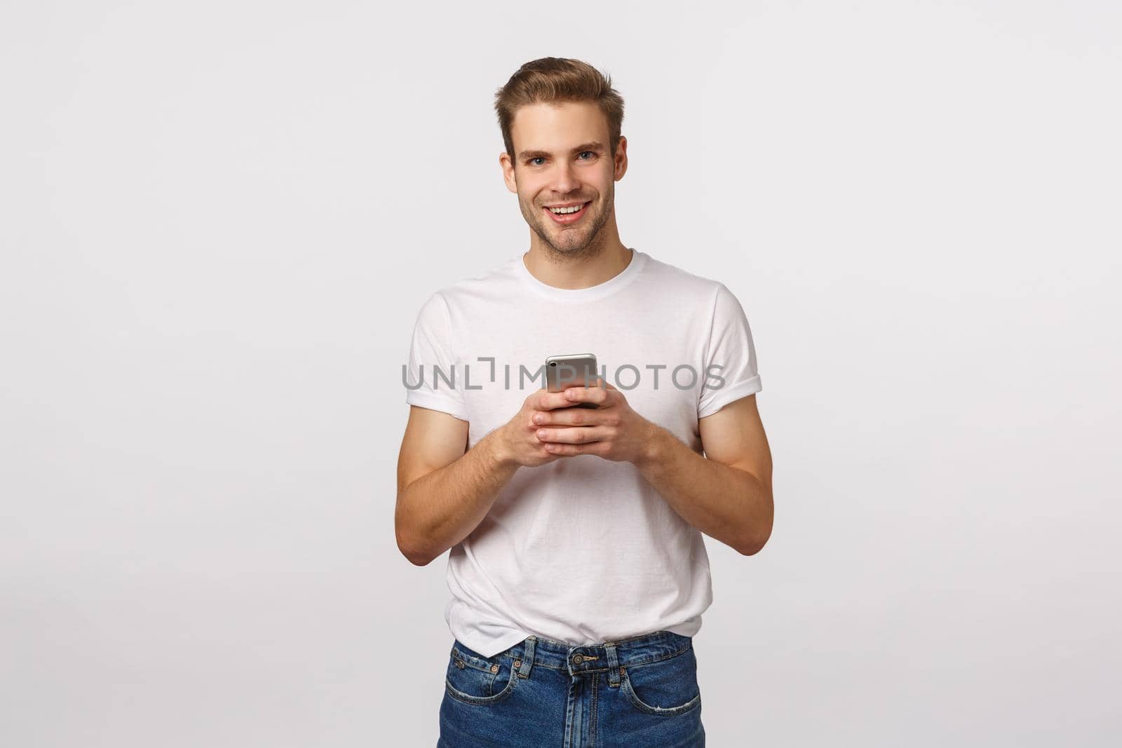 Telephone, digital modern life, technology concept. Handsome smiling blond caucasian man with bristle, wearing white t-shirt and jeans, holding smartphone as chatting in messanger, look camera.