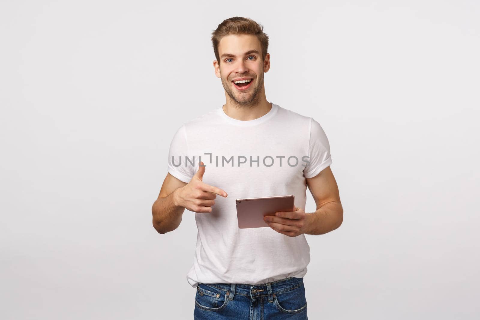 Smiling blond caucasian coworker male, wear t-shirt, jeans, hold digital tablet and pointing device, gadget display, grinning fascinated, found excellent website, good application, try new filters.