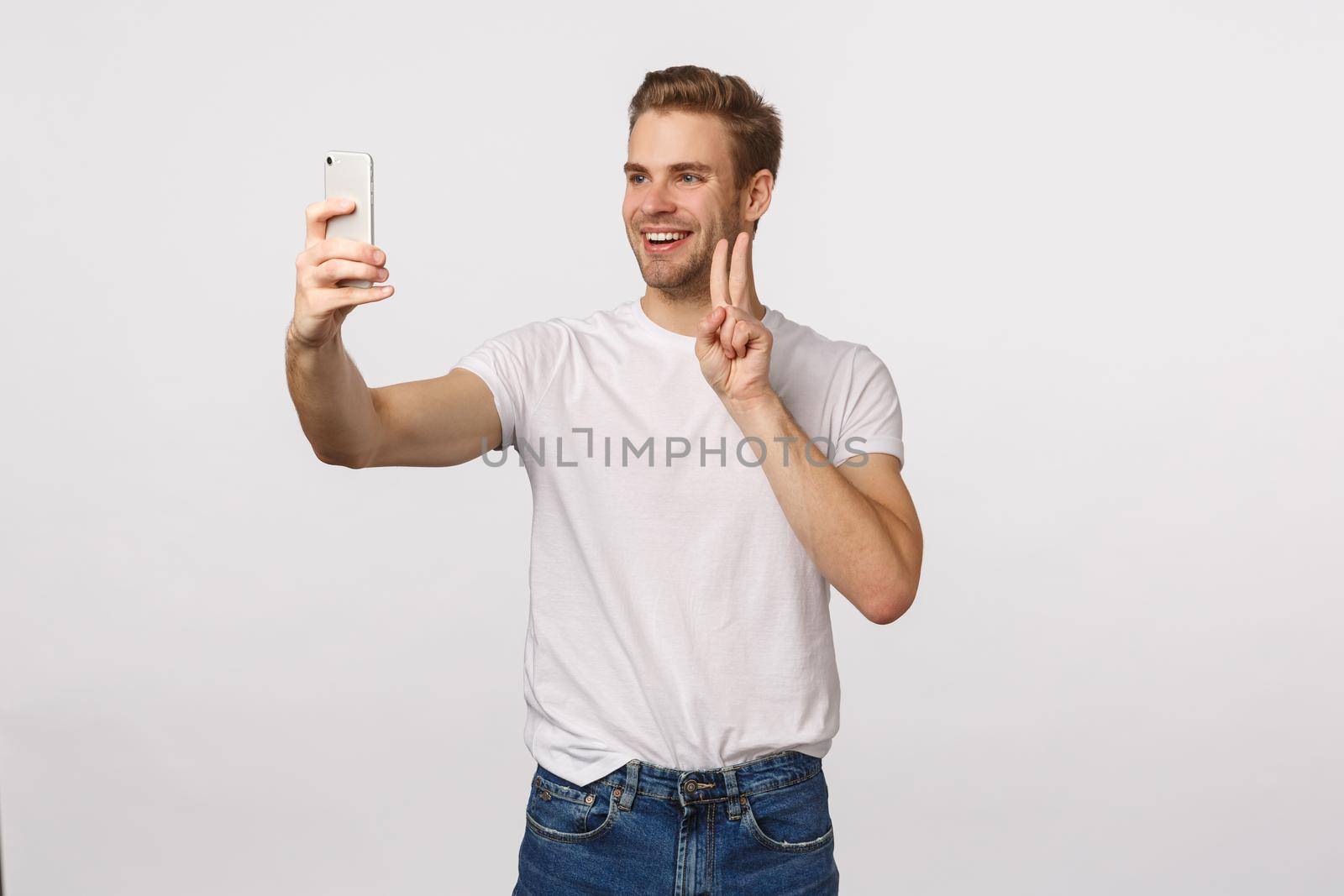 Cheerful man download new app, trying out cute filters as taking selfie on smartphone, making peace sign and smiling joyfully, photographing himself, standing white background.