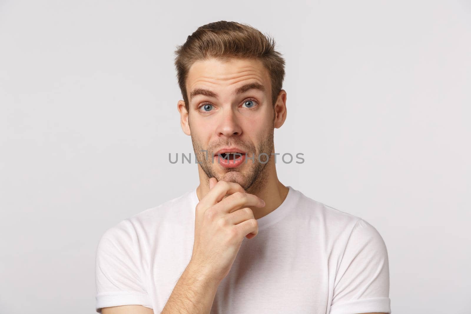 Impressed and surprised, curious good-looking blond bearded male in white t-shirt, touchign chin, open mouth and stare astonished, listen interesting suggestion, stand white background.