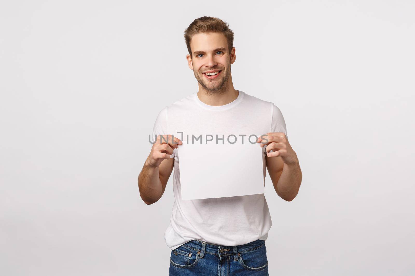 Attractive blond bearded handsome man in white t-shirt, holding blank empty paper as promoting corporate banner, promo, website or advertisement, smiling pleased, white background.