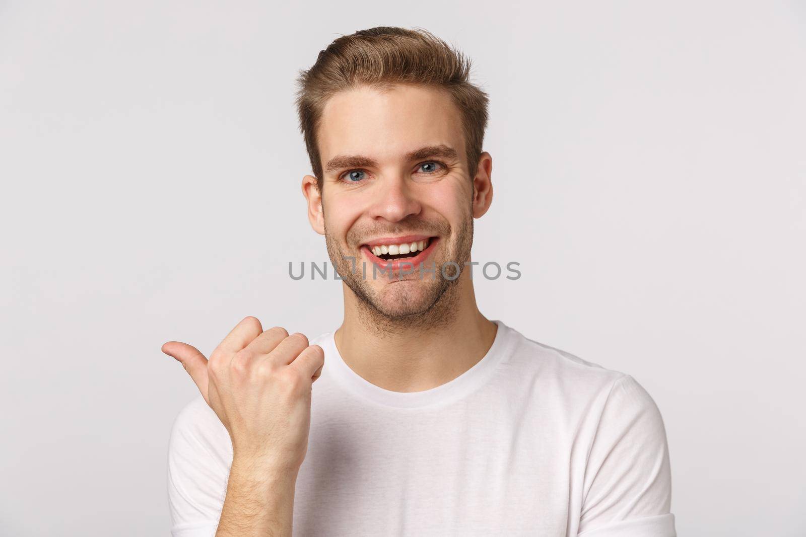 Close-up studio portrait blond handsome and joyful, smiling man asking friend opinion as pointing thumb left, discuss company product, grinning cheerful, standing white background.