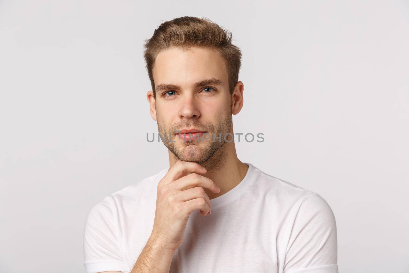 Hmm let me think. Pensive good-looking blond young bearded man in white t-shirt, touching chin and squinting intrigued, thinking, pondering what choice make, standing thoughtful.