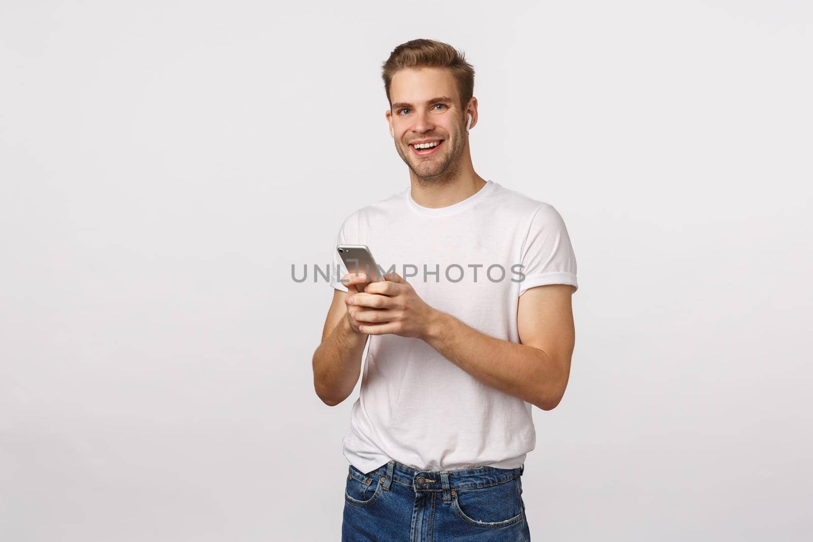 Joyful charming, smiling caucasian blond male in white t-shirt, listen music wireless headphones, holding smartphone, talking to girlfriend using earphones, picking song, make playlist.