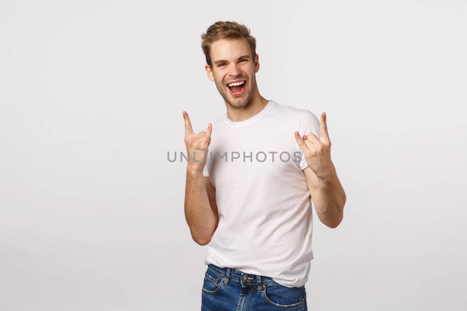 This party rocks my world. Excited and carefree, happy charming blond bearded man in white t-shirt, show heavy metal, rock-n-roll gesture and smiling, attend fantastic festival, enjoy event.