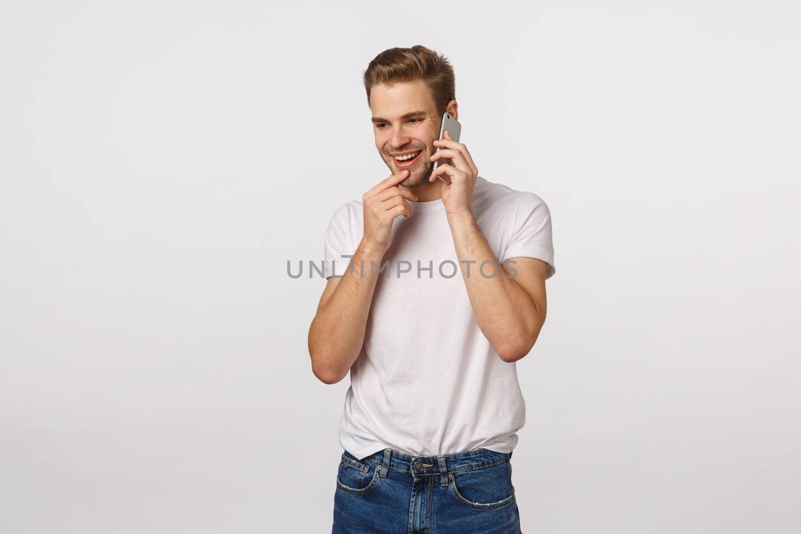 Intrigued and happy excited blond guy receive invitation party via phone call, holding smartphone near ear, talking on mobile, touching lip and smiling tempting, have conversation, white background.