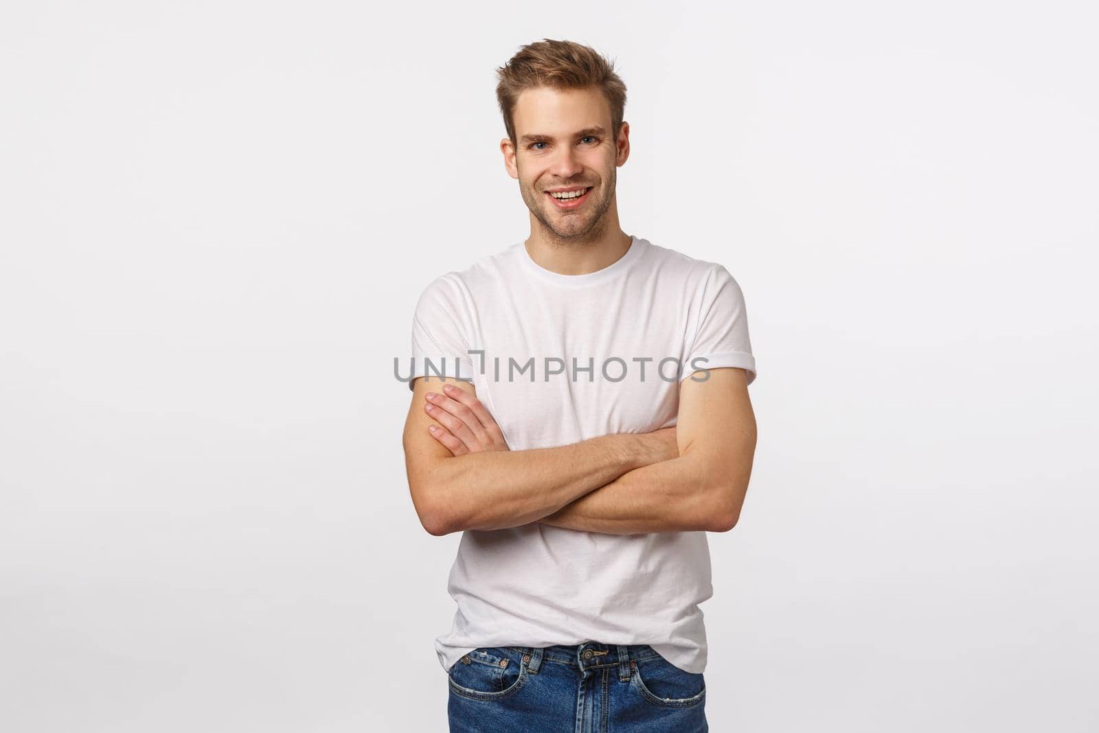 Enthusiastic, happy smiling blond man with bristle, white t-shirt, jeans, cross arms chest and grinning amused, hear interesting story, accepting good idea, agree with person, white background.