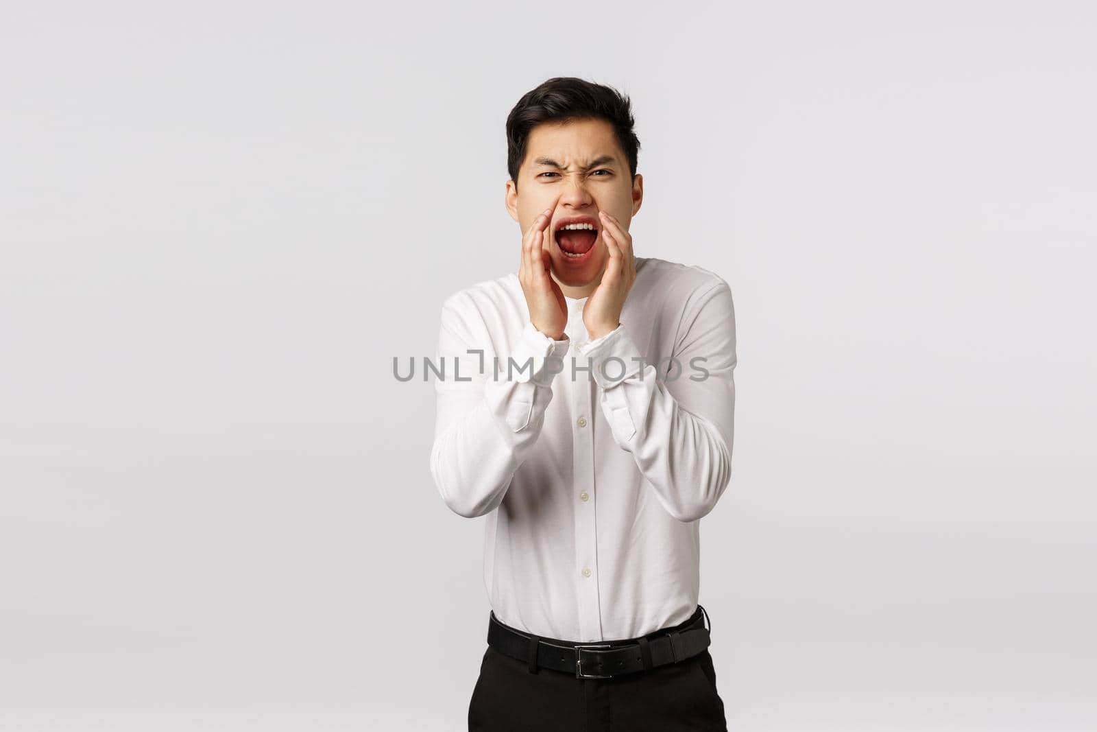 Guy swearing and yelling out loud cursing words. Angry, outraged and displeased asian man hold hands near opened mouth like megaphone, shouting at someone arguing or having fight, white background.