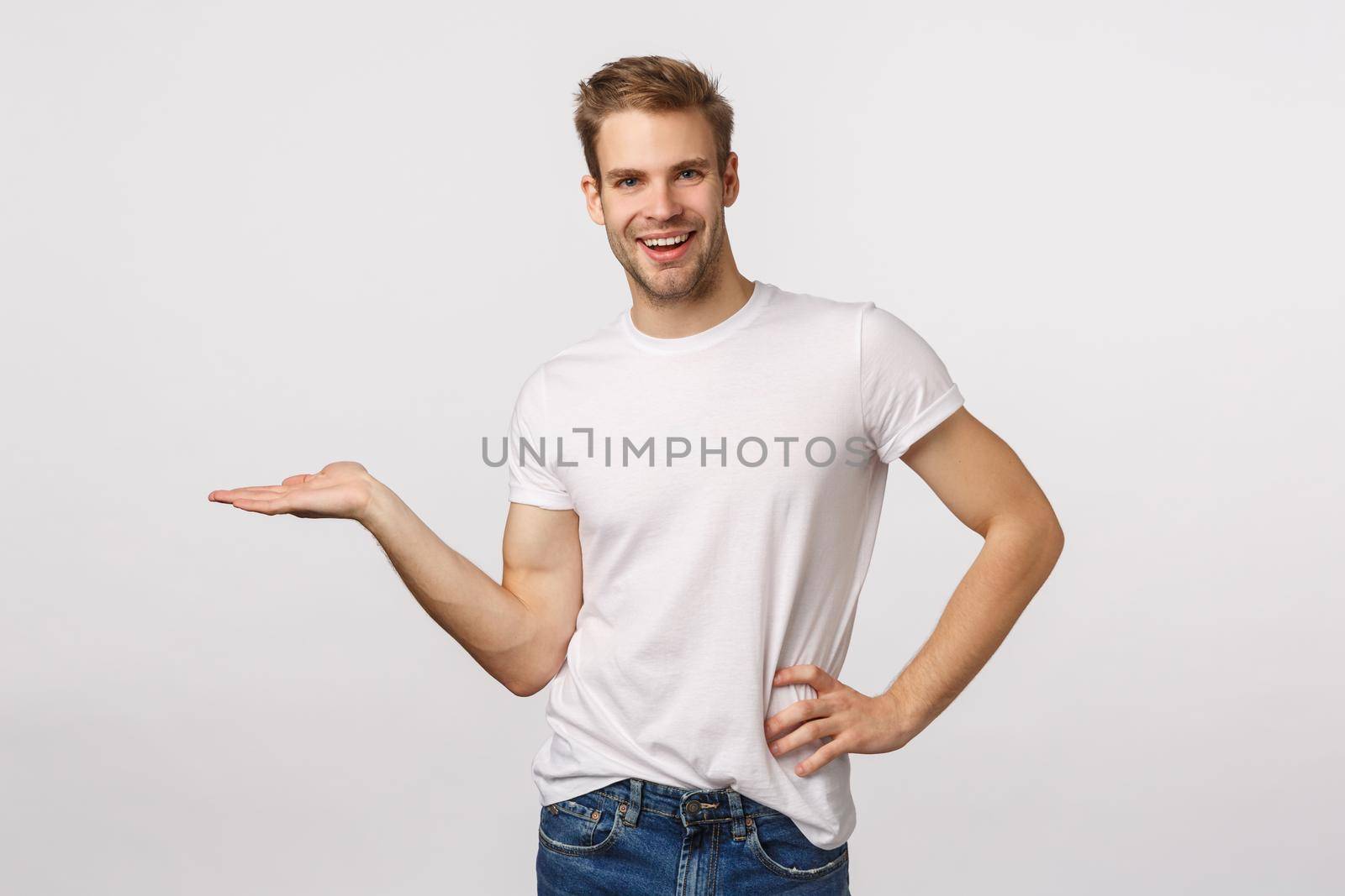 Cute smiling, happy young blond bearded man in white t-shirt introduce product, holding something on blank copy space, introduce chart or concept, grinning delighted, white background.