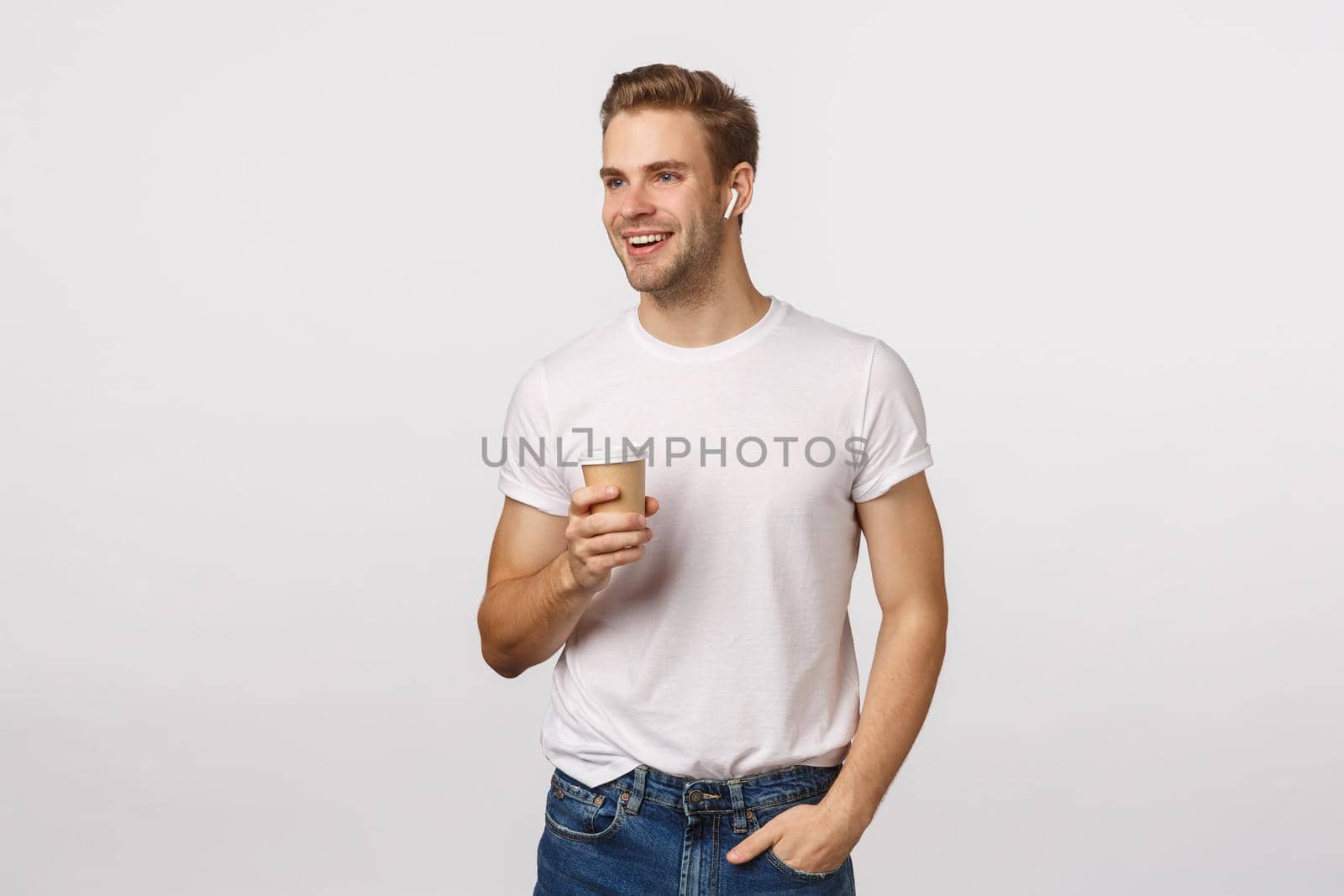 Modern life, lifestyle and people concept. Attractive blond caucasian guy holding take-away paper cup, enjoy coffee and talking phone via wireless headphones, smiling look away, white background.