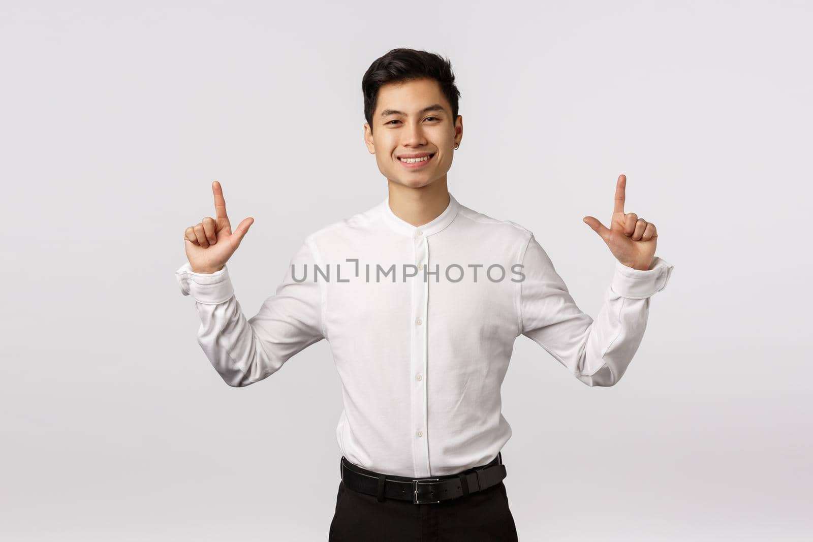 Hansdome asian businessman in white shirt, black pants, pointing fingers up and smiling with pleased, satisfied or glad expression, showing product, promoting corporate banner, white background.