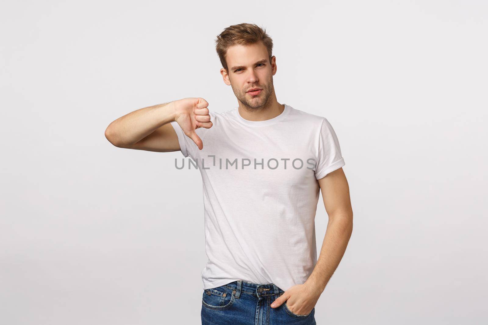 Serious-looking, skeptical and judgemental handsome blond bearded guy show thumb-down and looking disappointed, give negative feedback, upset with poor results, judging, white background.