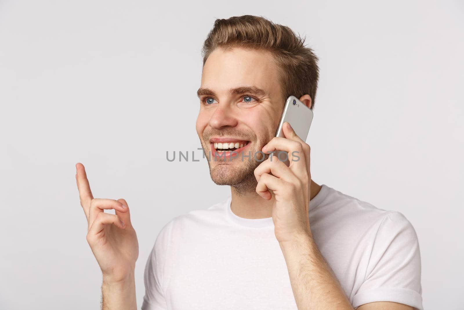 Close-up blond handsome man with blue eyes, white t-shirt, holding smartphone near ear, gesturing finger, smiling pleased, make order, confirm online shopping, talking with friend, white background.