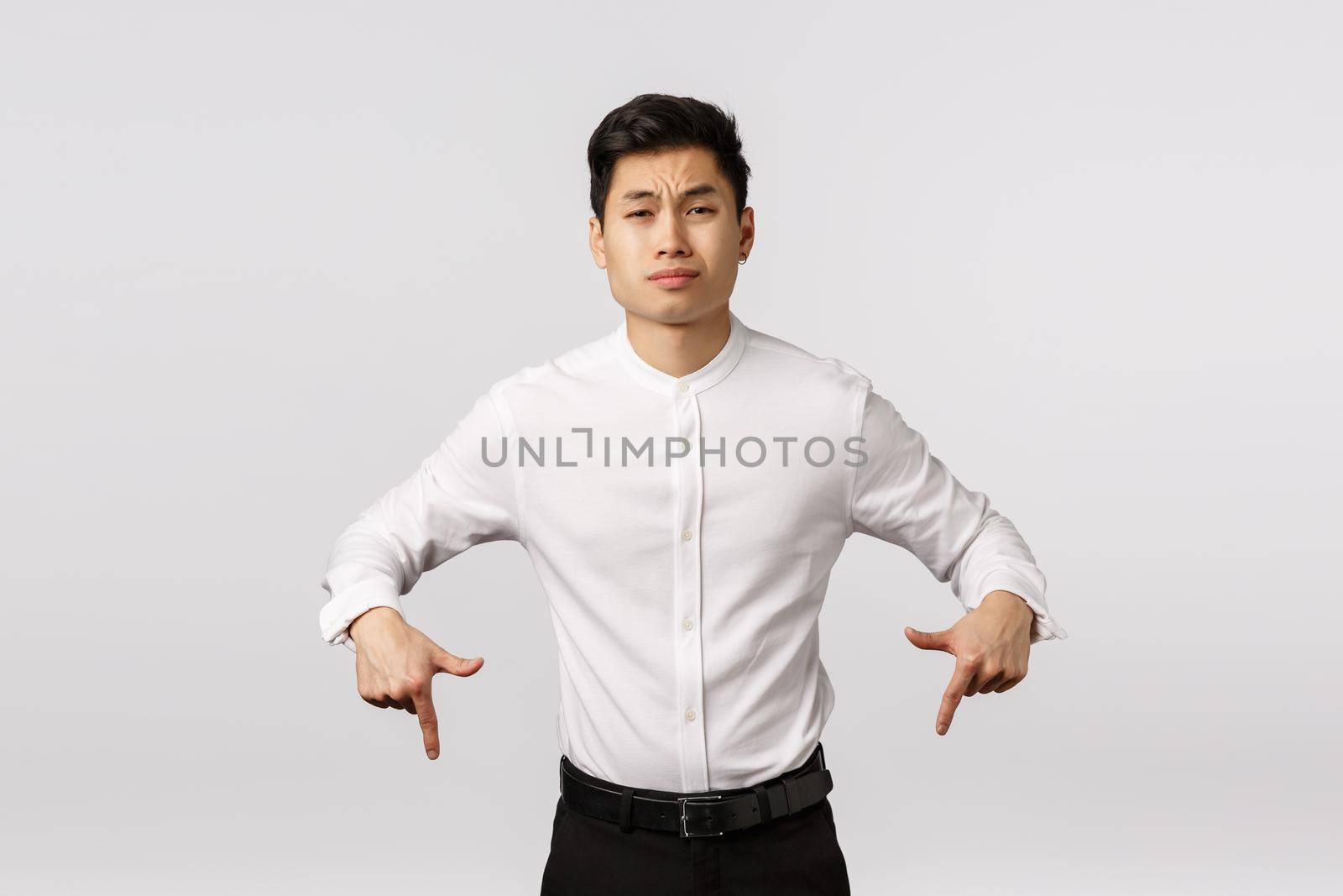 Skeptical arrogant asian businessman in white shirt, pants, pointing down look camera unsatisfied and unimpressed, sulking, grimacing careless, express dislike or scorn, white background.
