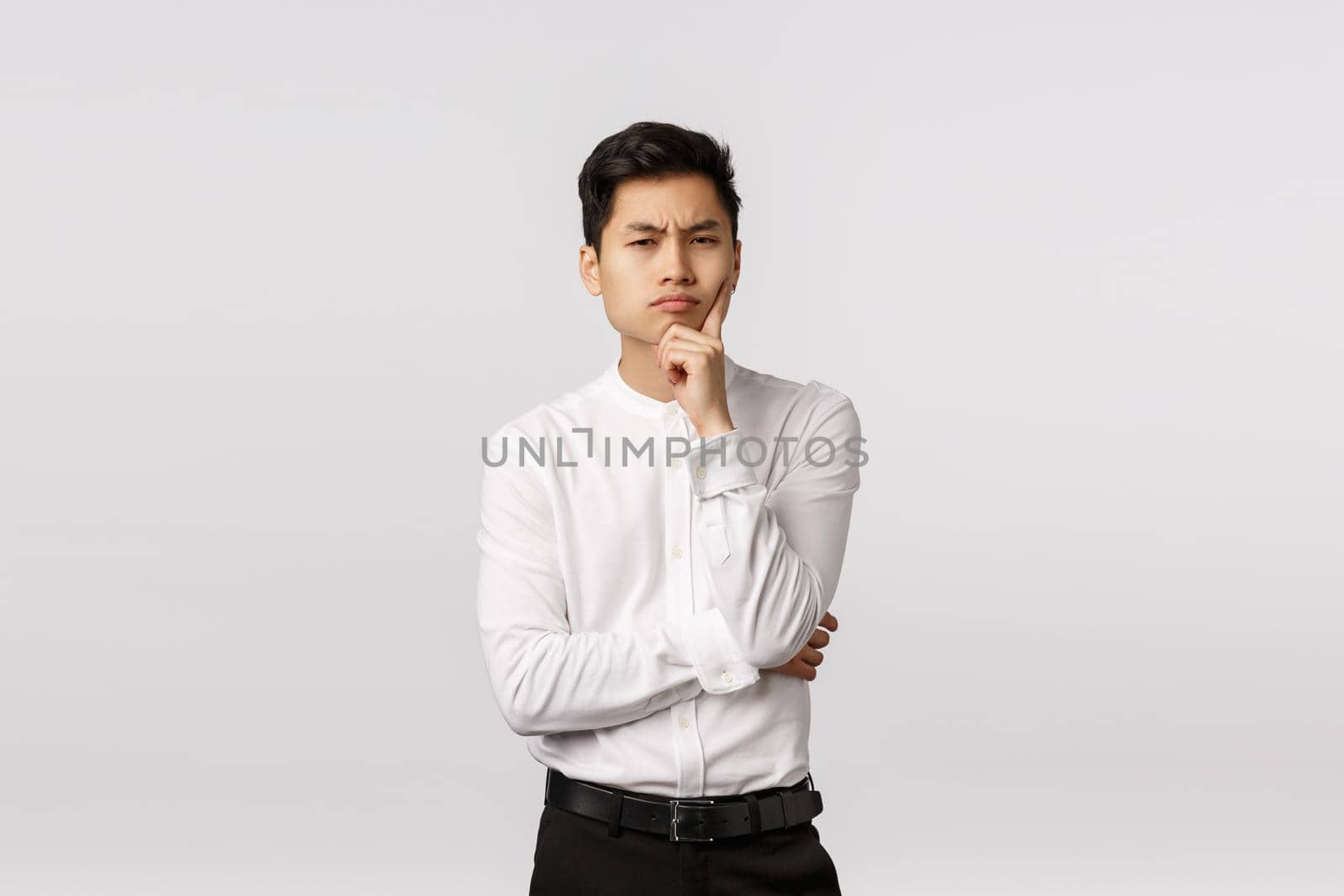 Suspicious and skeptical, thoughtful attractive asian man in shirt, pants, frowning look camera with disbelief and serious face, touch chin, thinking, have assumptions, standing white background.