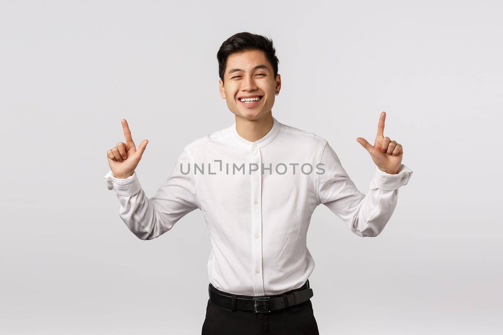 Happiness, wellbeing and success concept. Cheerful cute asian guy in white shirt, black pants, smiling and laughing happily, sharing positive emotions and pointing up at product banner.