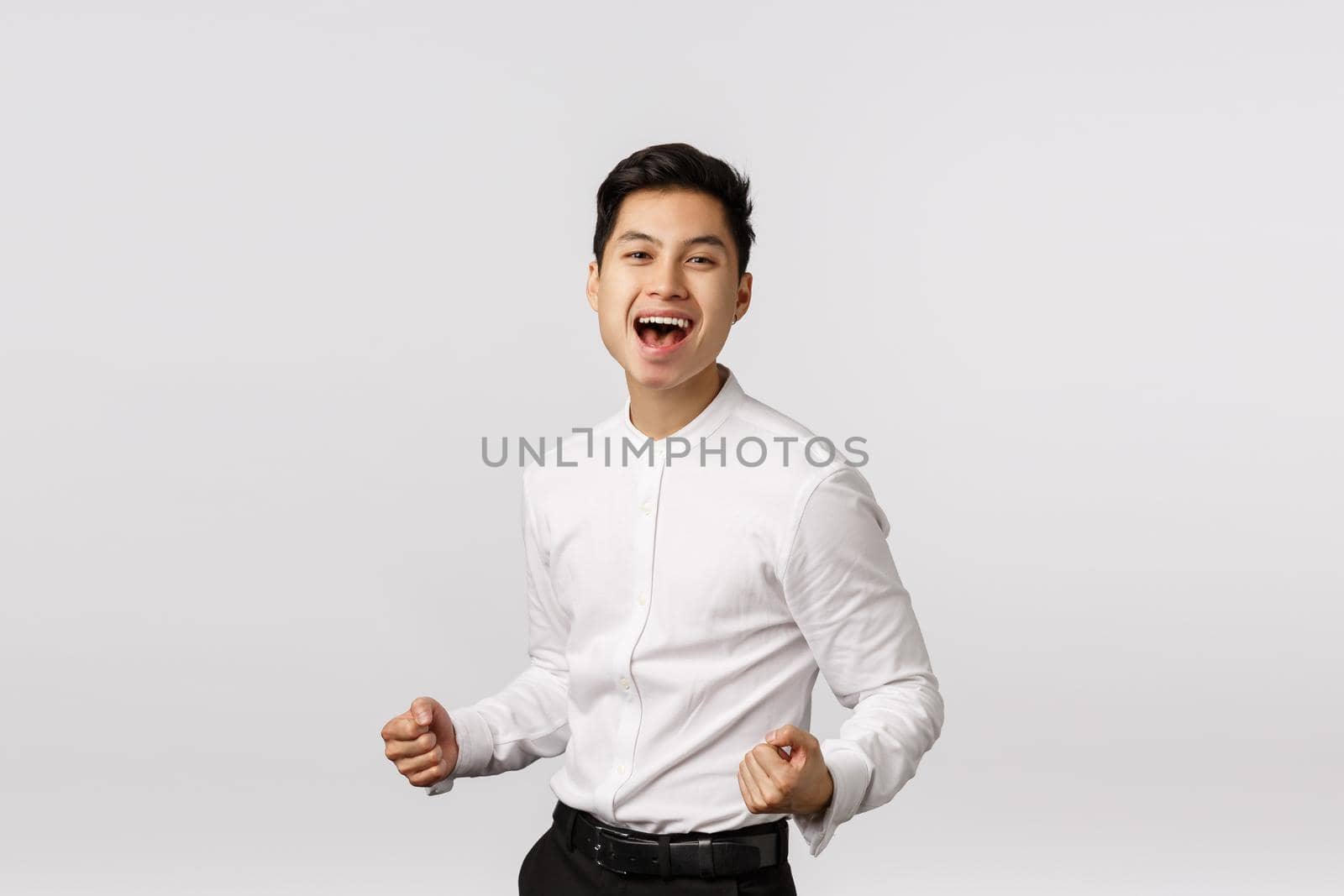 Delighted, successful young asian businessman clench fists, pump it up and smiling pleased, say yes, celebrating win, placed good bid, achieve goal, rejoicing receive prize, standing white background.