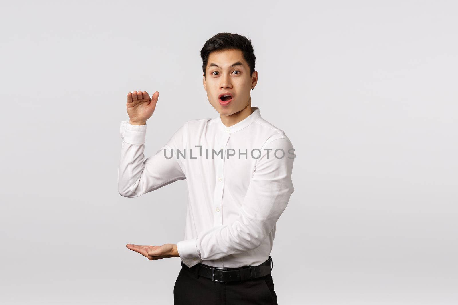 Impressed lucky handsome asian man in white shirt, pants, open mouth say wow astonished, showing big box, large product with hands gesture, standing fascinated white background.