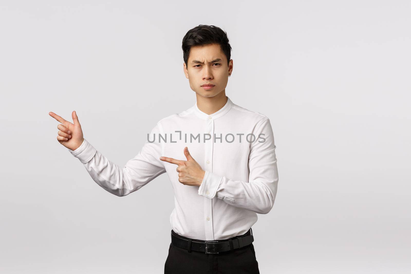 Serious-looking strict and unsatisfied young asian man disappointed with painting hanging wrong. Businessman looking camera sulking and frowning displeased, pointing left to turn attention at mistake by Benzoix