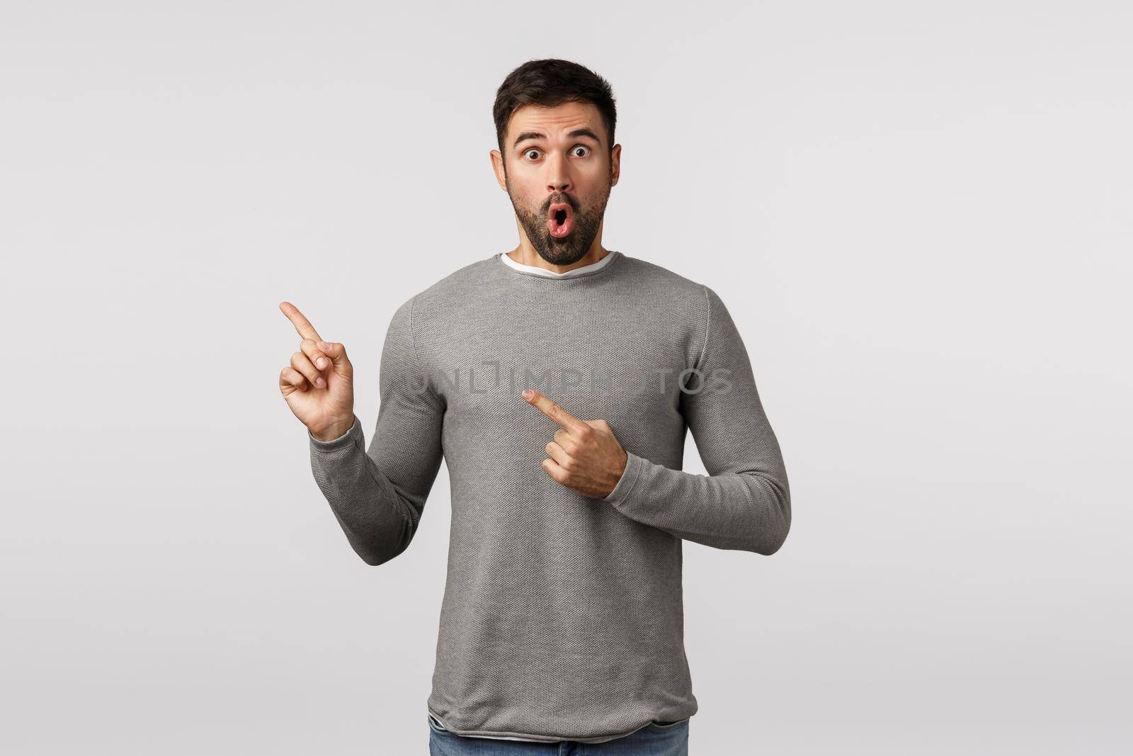 Wow amazing surprise, look at that. Amazed and astonished handsome bearded man in grey sweater, gasping wondered, stare impressed, pointing upper left corner, indicate promo, white background by Benzoix
