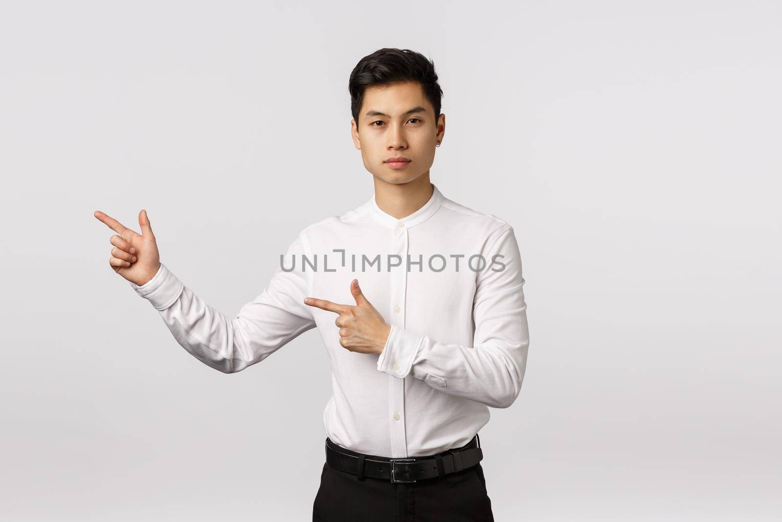 Serious-looking asian businessman give direction, pointing left, recommend visit site link, look corporate banner. Attractive elegant young man in white shirt and black pants advertising product.