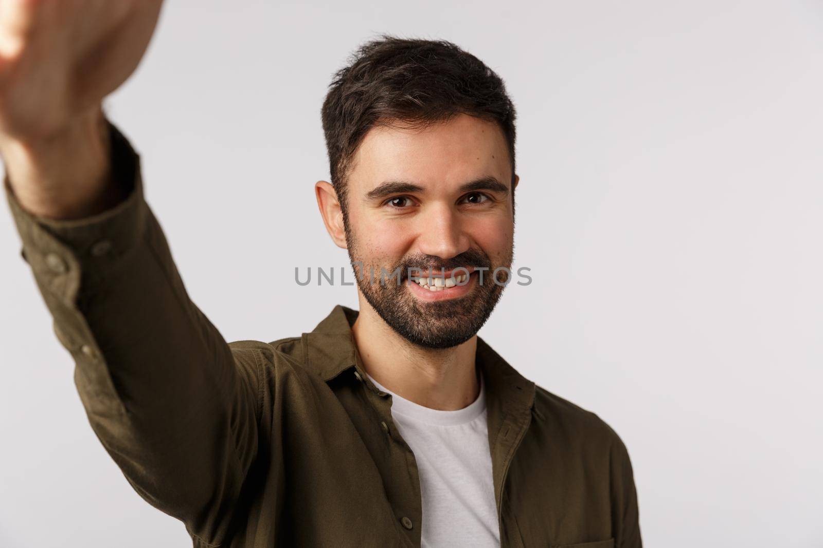 Handsome cheerful bearded man in coat want upload new photo gay dating application, hold smartphone with arm and smiling camera, taking selfie, standing white background joyful. Copy space