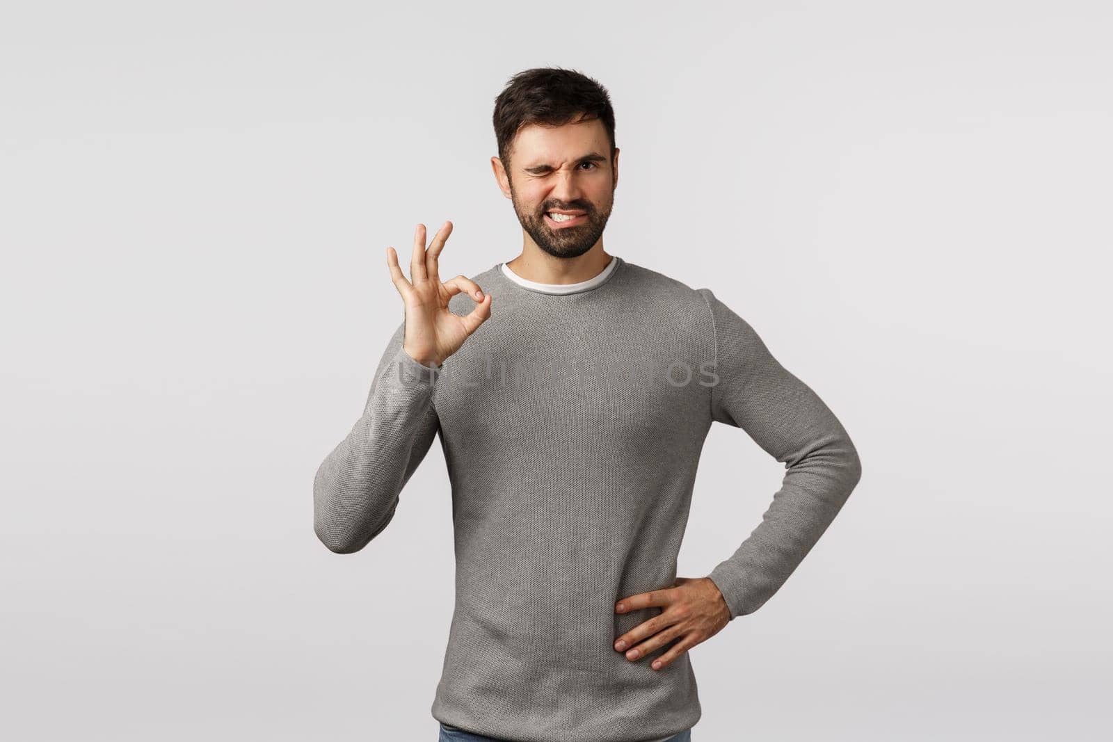 Everything under control. Sassy, assertive charismatic bearded man in grey sweater, wink confident, cant rely on me, show okay, ok gesture, approve product, like idea, accept plan, white background.