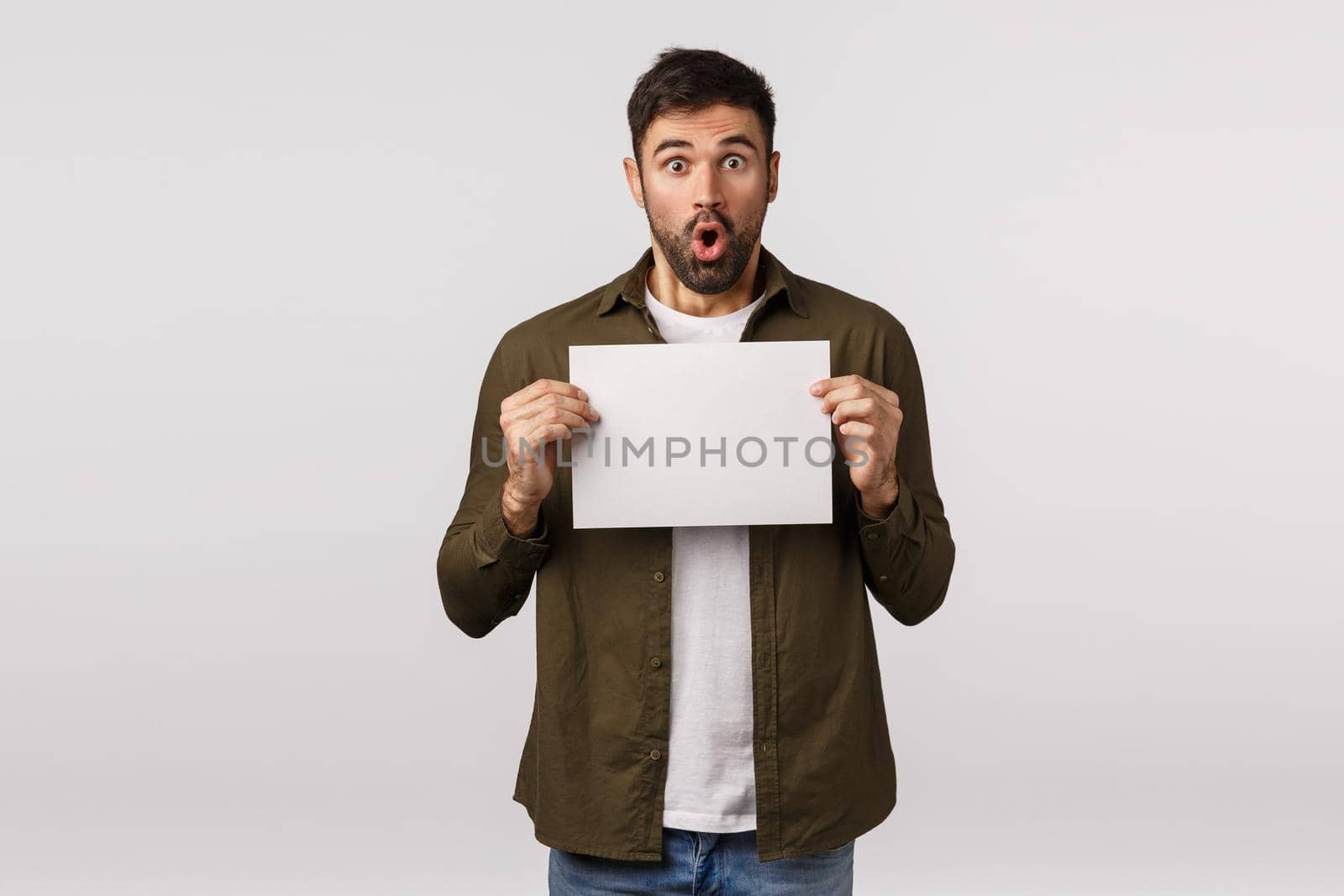 Amused, surprised handsome bearded guy holding blank paper near chest and folding lips in awe, fascinated, write good deal, promo offer, recommend visit shopping site, white background.