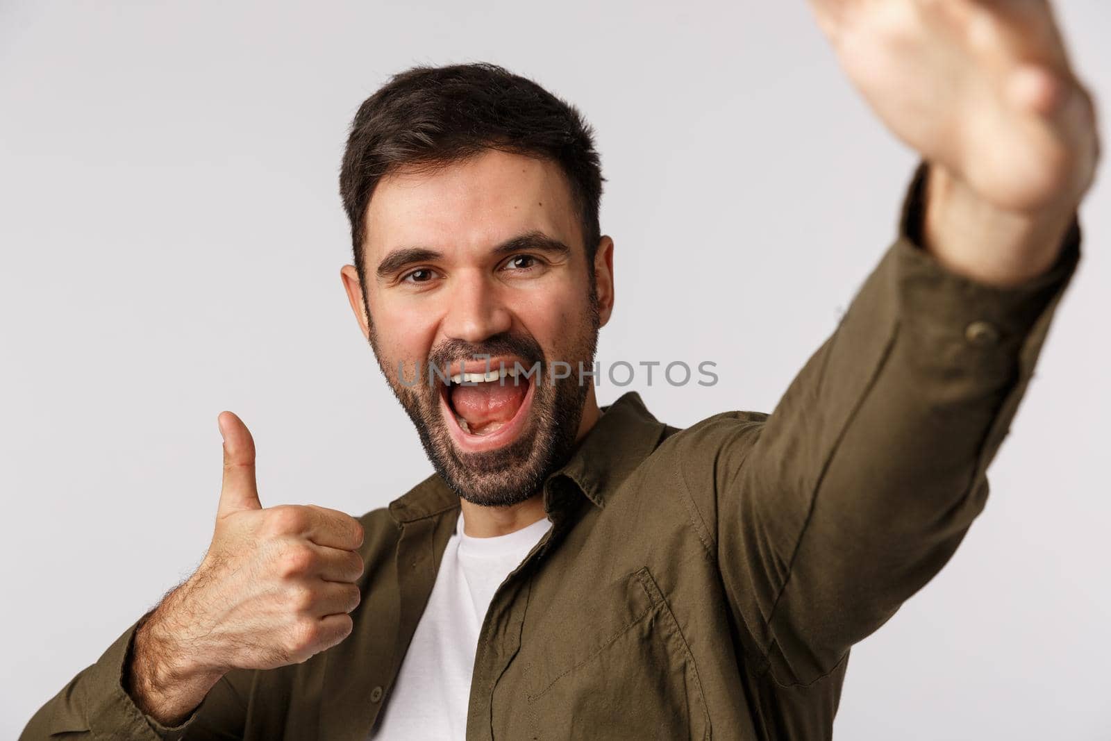 Excited, upbeat handsome bearded man absolutely adore party, like event, hold smartphone with one arm and show thumb-up, taking selfie, record vlog, online stream, standing white background by Benzoix