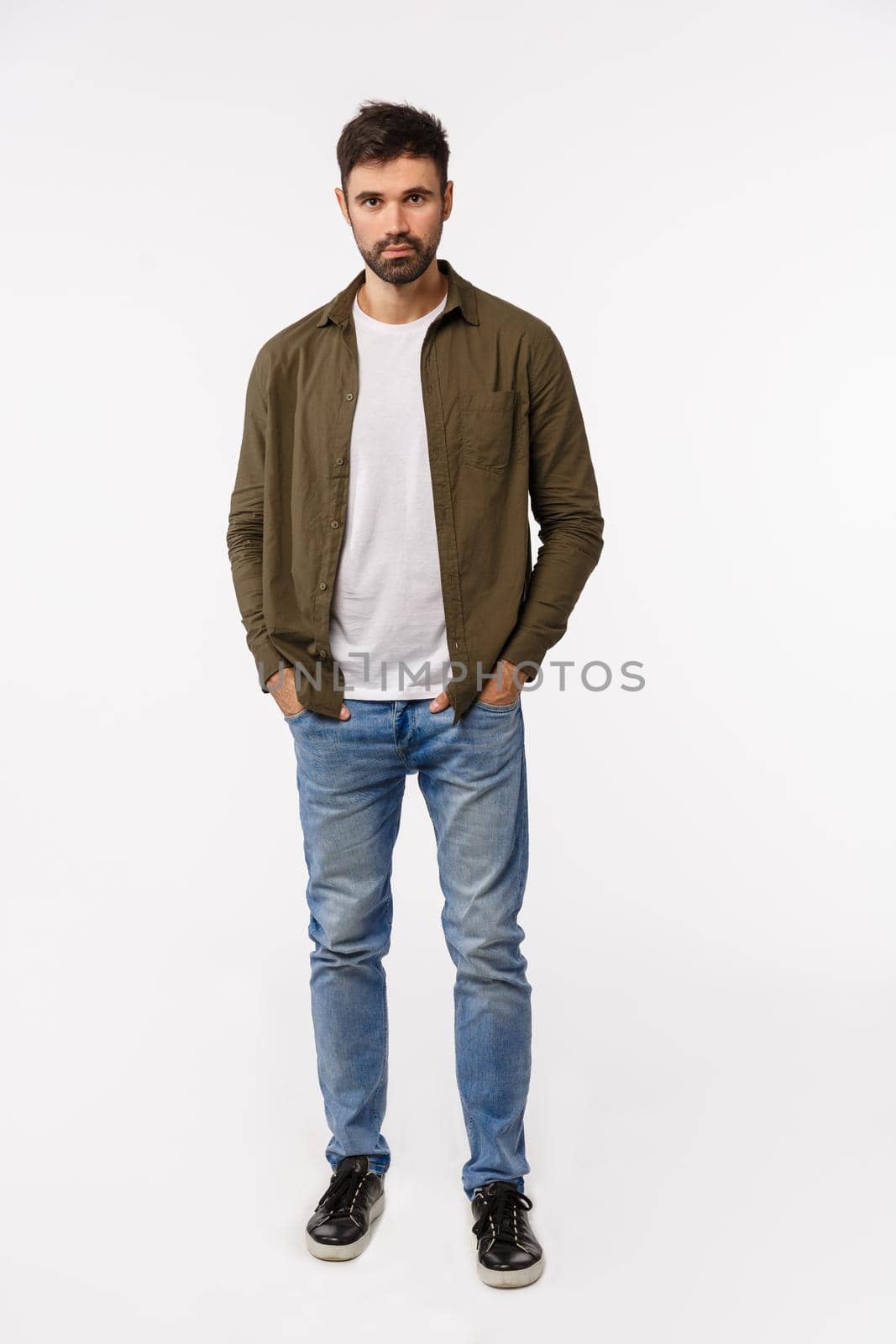 Vertical full-length shot sassy and confident modern man in coat, jeans streetstyle outfit, holding hands in pockets, looking assertive, smiling, getting ready business appointment, white background.