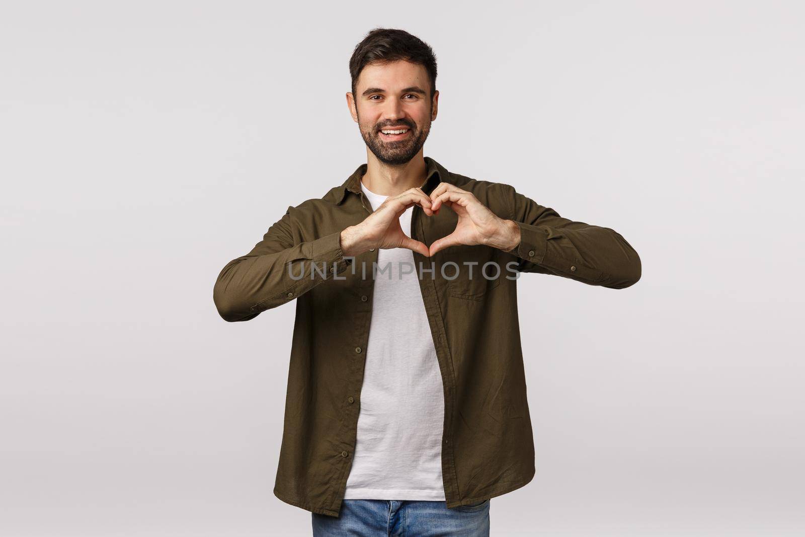 Valentines day, affection and relationship concept. Cheerful tender bearded male in coat, showing heart gesture near chest, express love or adornment, smiling cute, express gratitude or affection by Benzoix