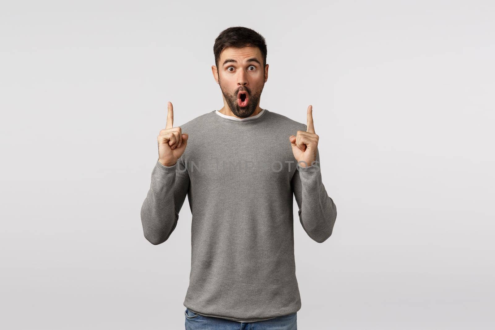 Surprised and amazed, speechless excited handsome bearded guy in grey sweater, say wow, folding lips amused, gasping astonished, pointing fingers up to turn attention, discuss impressive offer.