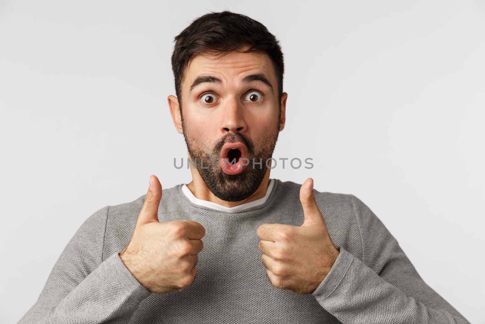 Wonderful idea, totally approve. Amazed and fascinated handsome bearded male in grey sweater, show thumbs-up and gasping in amazement and awe, folding lips say wow stare camera, white background.