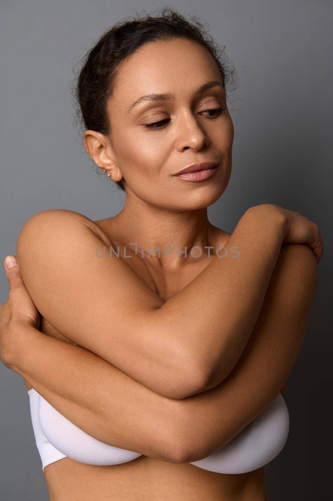 Close-up beauty portrait of self-confident middle aged African woman with perfect clean shiny healthy skin, hugging herself, isolated over gray background with copy space. Body and skin care concept by artgf