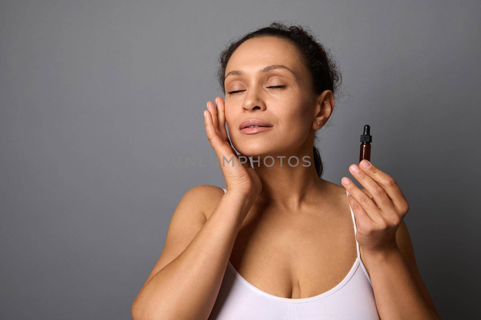 Beautiful mature woman enjoys doing facial massage with beauty product, poses with closed eyes on gray background with copy space for spa salons, aesthetic medicine clinics ad. Body skin care concept