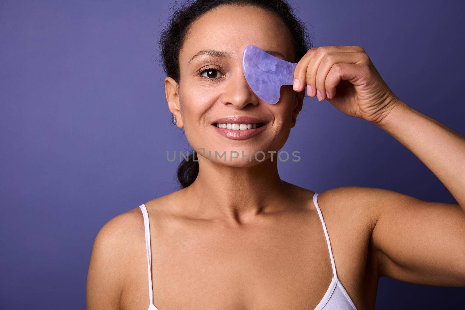 Beautiful half-naked African woman smiles toothy smile, holds Ayurvedic Gua Sha jade stone massager near her face, looking at the camera, posing on purple background with a copy space for advertising by artgf