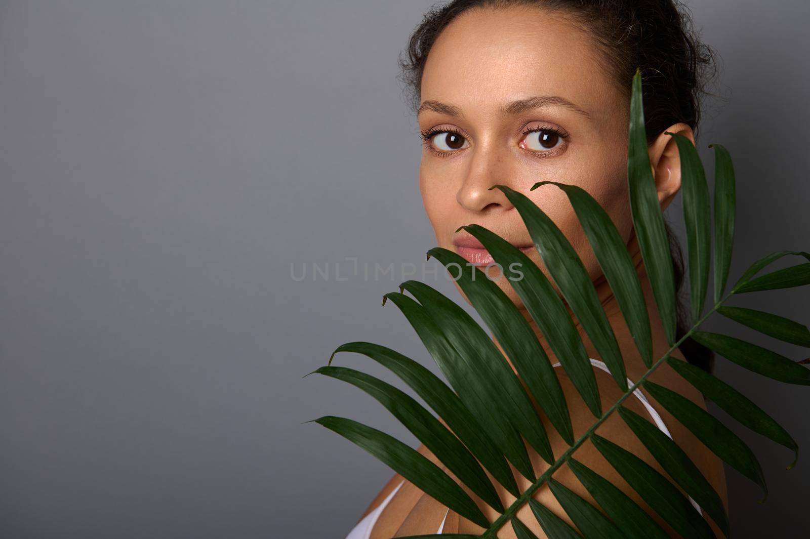 Beautiful woman with natural makeup and clean fresh healthy skin covers her face with palm leaf, looking confidently at camera, posing against gray wall background with copy space for ad. Spa concept