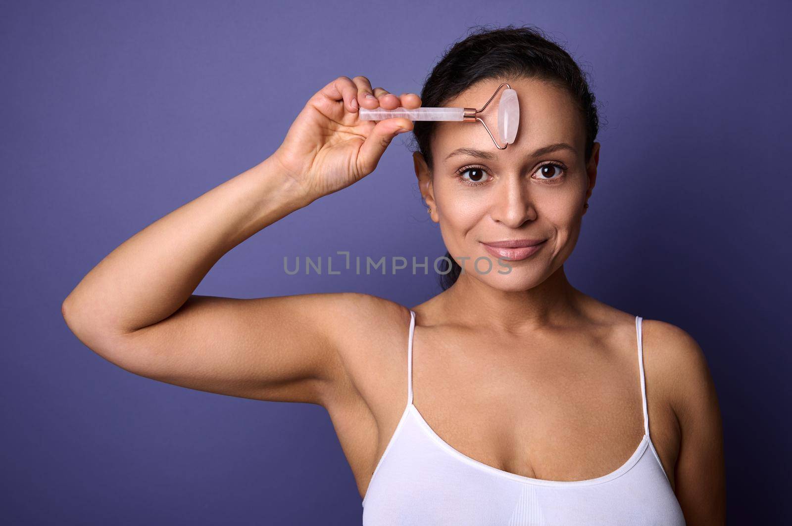 Beautiful Hispanic middle aged woman in white underwear massaging her face with jade stone roller massager, doing smoothing facial massage, smiles posing against purple background with copy ad space by artgf