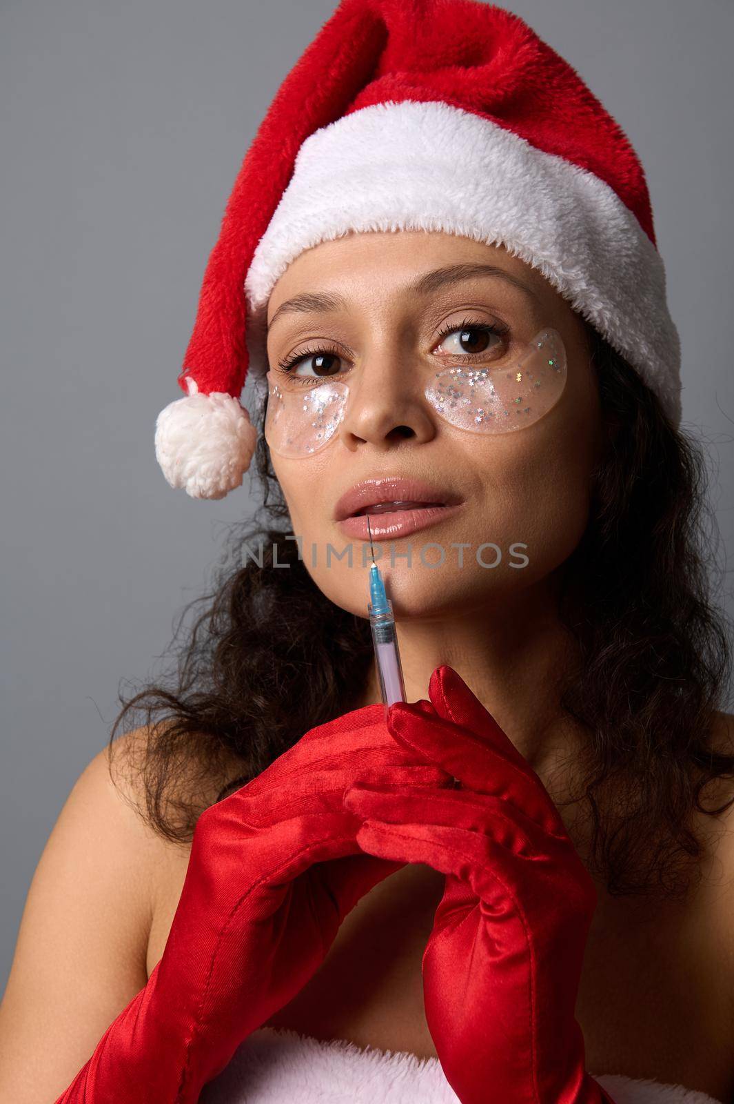 Headshot of beautiful woman with natural make-up and shiny eye patches under eyes, wearing Santa hat holds a syringe with injection near her face, cutely looks at camera. Injection cosmetology concept by artgf