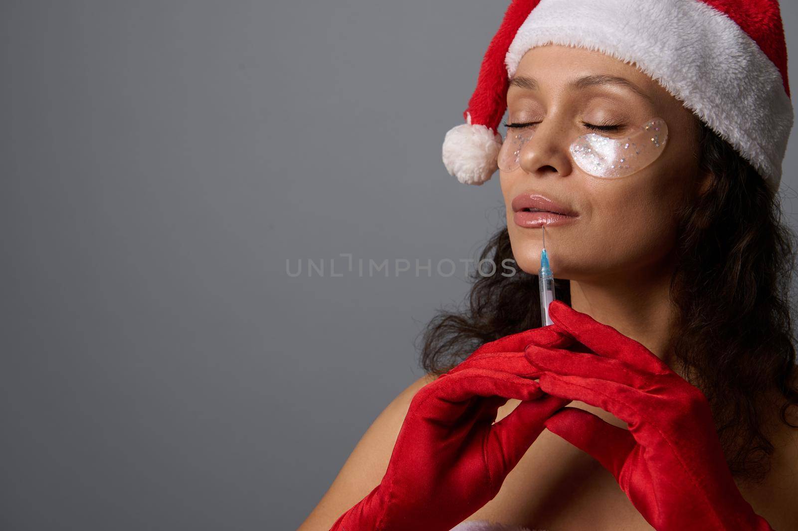 Beautiful woman dressed in Santa carnival attire, with patches under eyes receiving a beauty injection on her lips. Lips augmentation concept for Christmas advertising for aesthetic beauty salons. by artgf