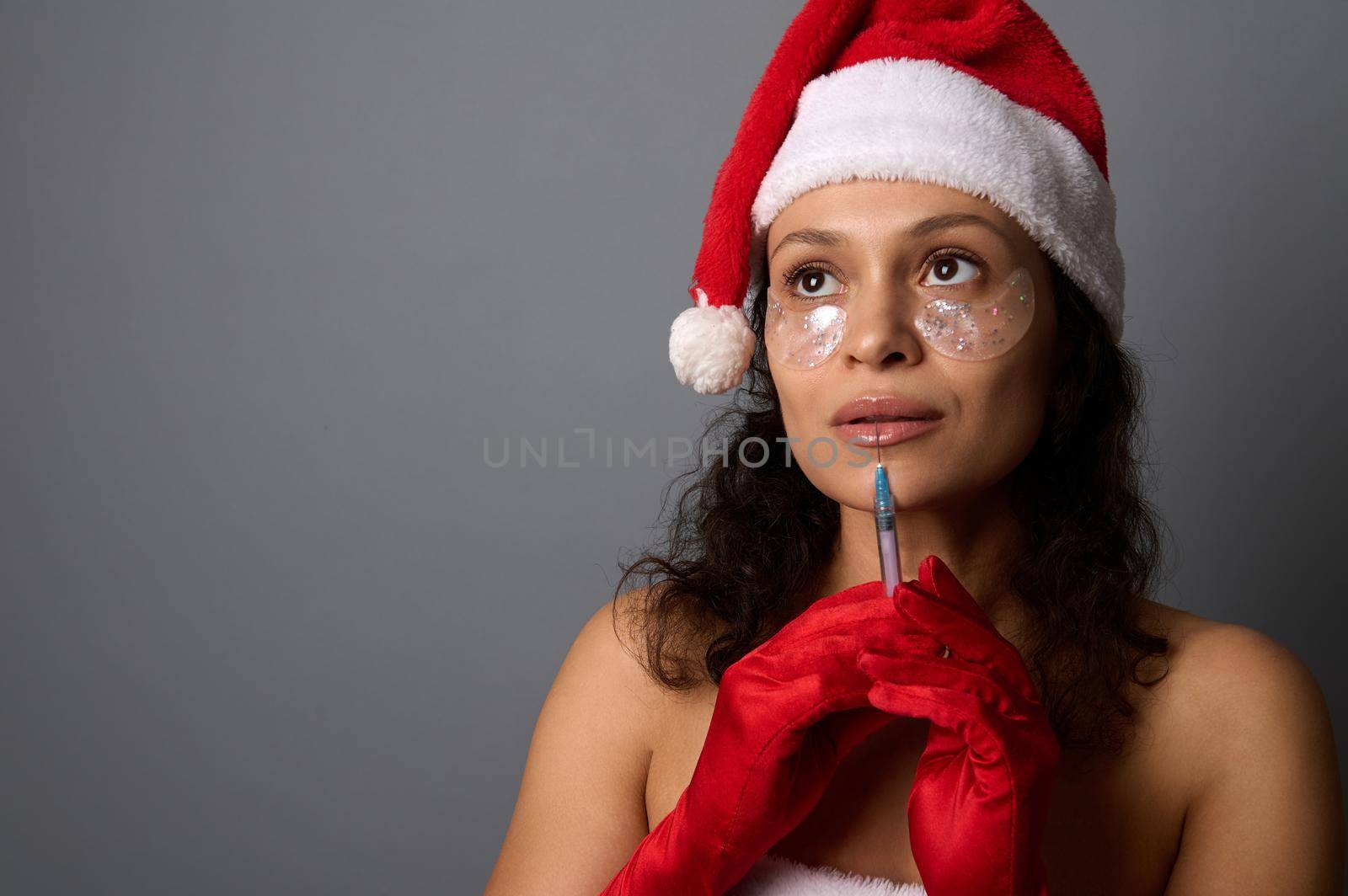 Pretty woman with natural make-up and patches under eyes, in Santa hat holds syringe with beauty injection near her face, looks up on copy space of gray background. Injection cosmetology, spa concept