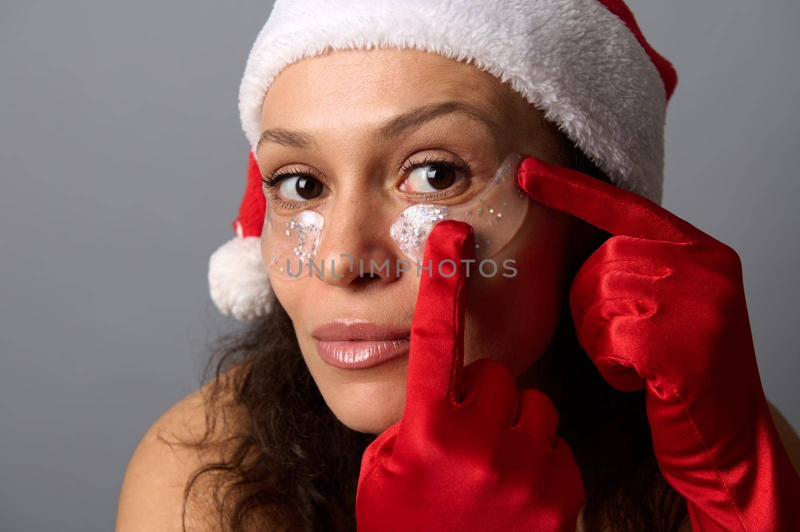 Close up face portrait of pretty brunette woman in Santa hat wearing eye patches looking closely at camera, isolated on gray background. Skin care concept for Christmas ad of cosmetology beauty salons