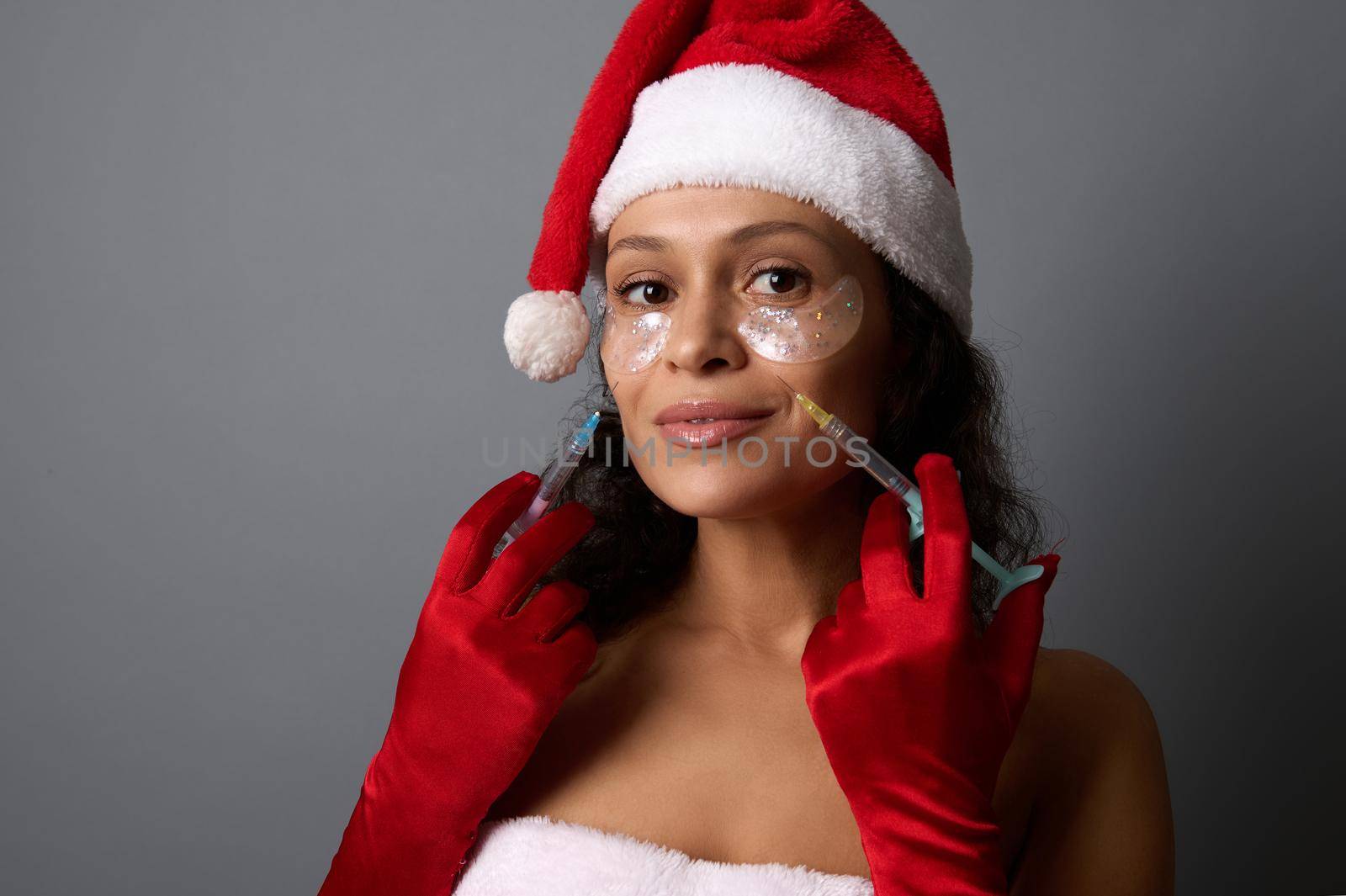 Beautiful woman dressed in Santa for Christmas holidays, holds syringes near her face, ready for beauty injections on her nasolabial folds. Anti-aging, face lifting and rejuvenation concept for ad by artgf