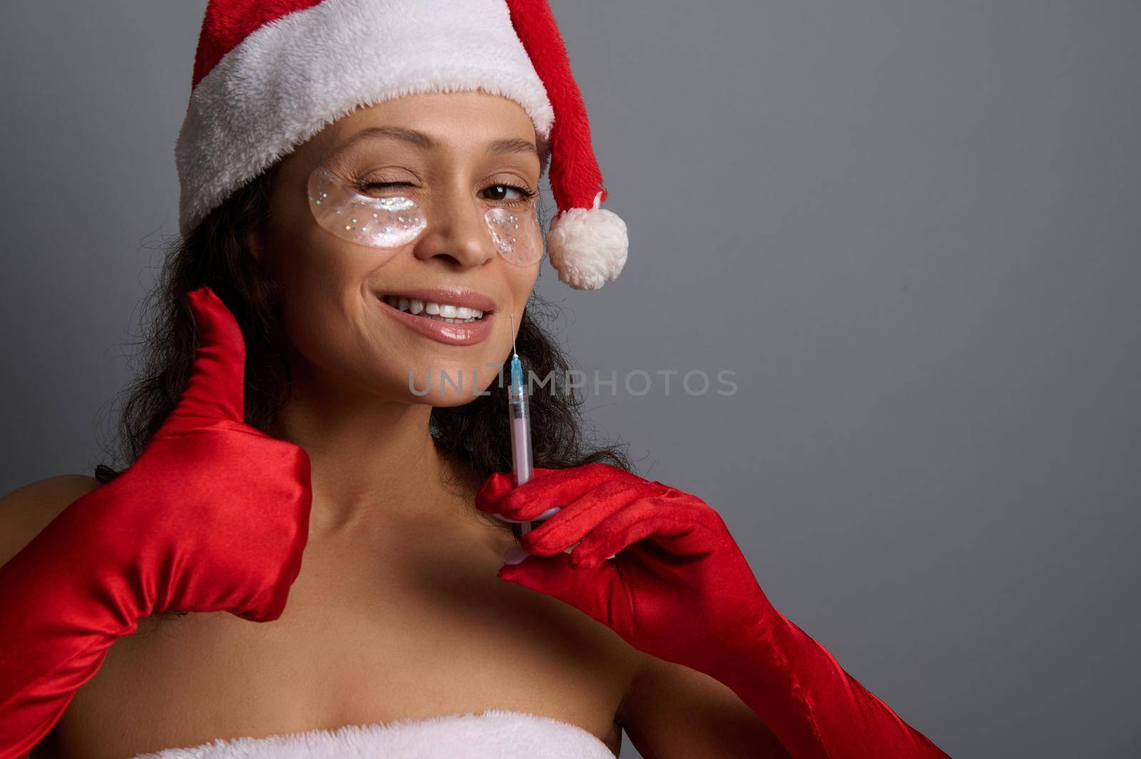 Sexy pretty woman with under eyes patches, dressed in Santa , holds syringe with beauty injection, shows thumb up, smiles to camera. Anti-aging, face lifting, rejuvenation, cosmetology concept for ad by artgf