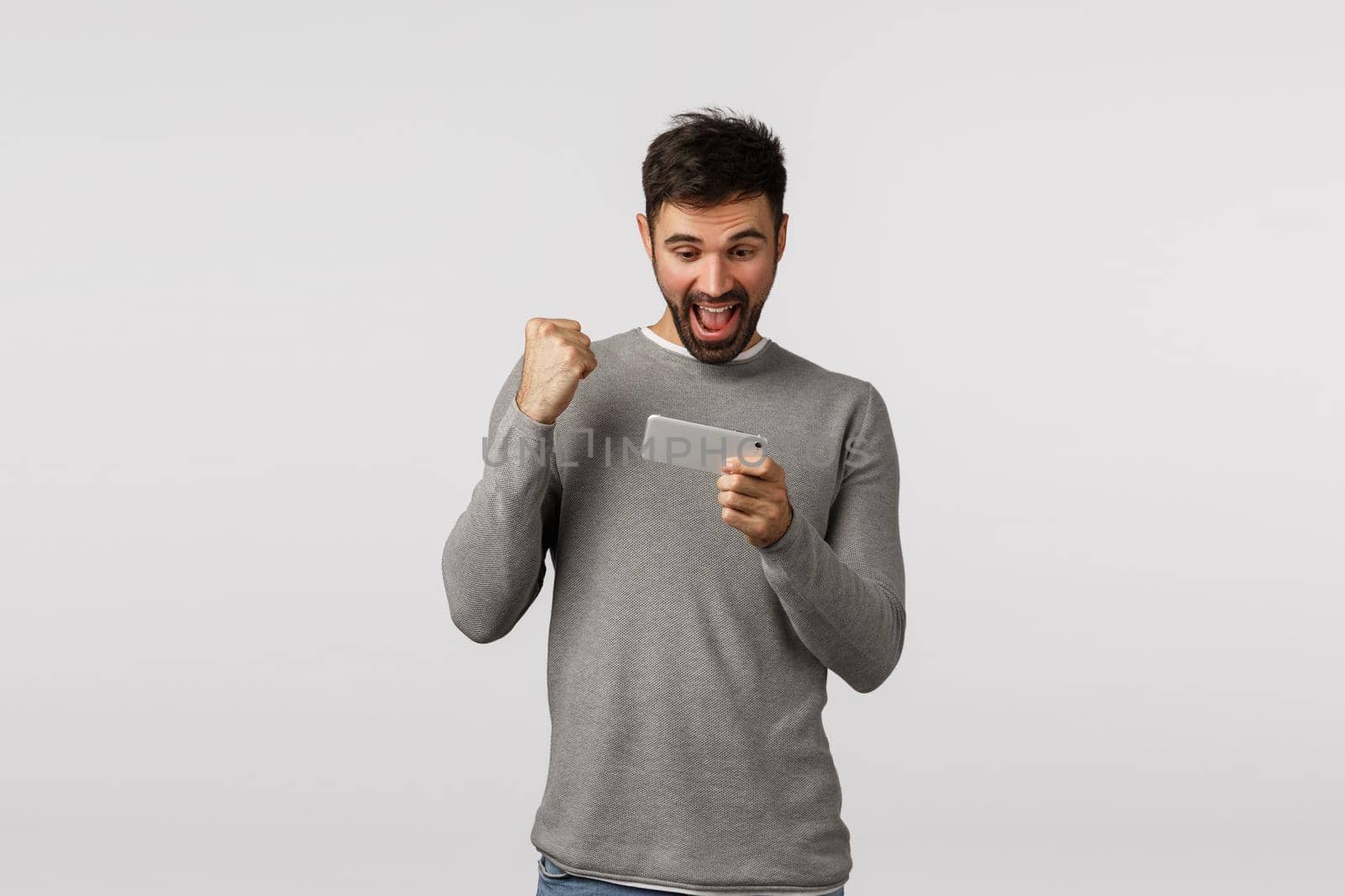 Happy and cheerful, excited bearded male in grey sweater, holding smartphone and fist pump as celebrating, say hooray or yes winning game level, see news in application, white background by Benzoix
