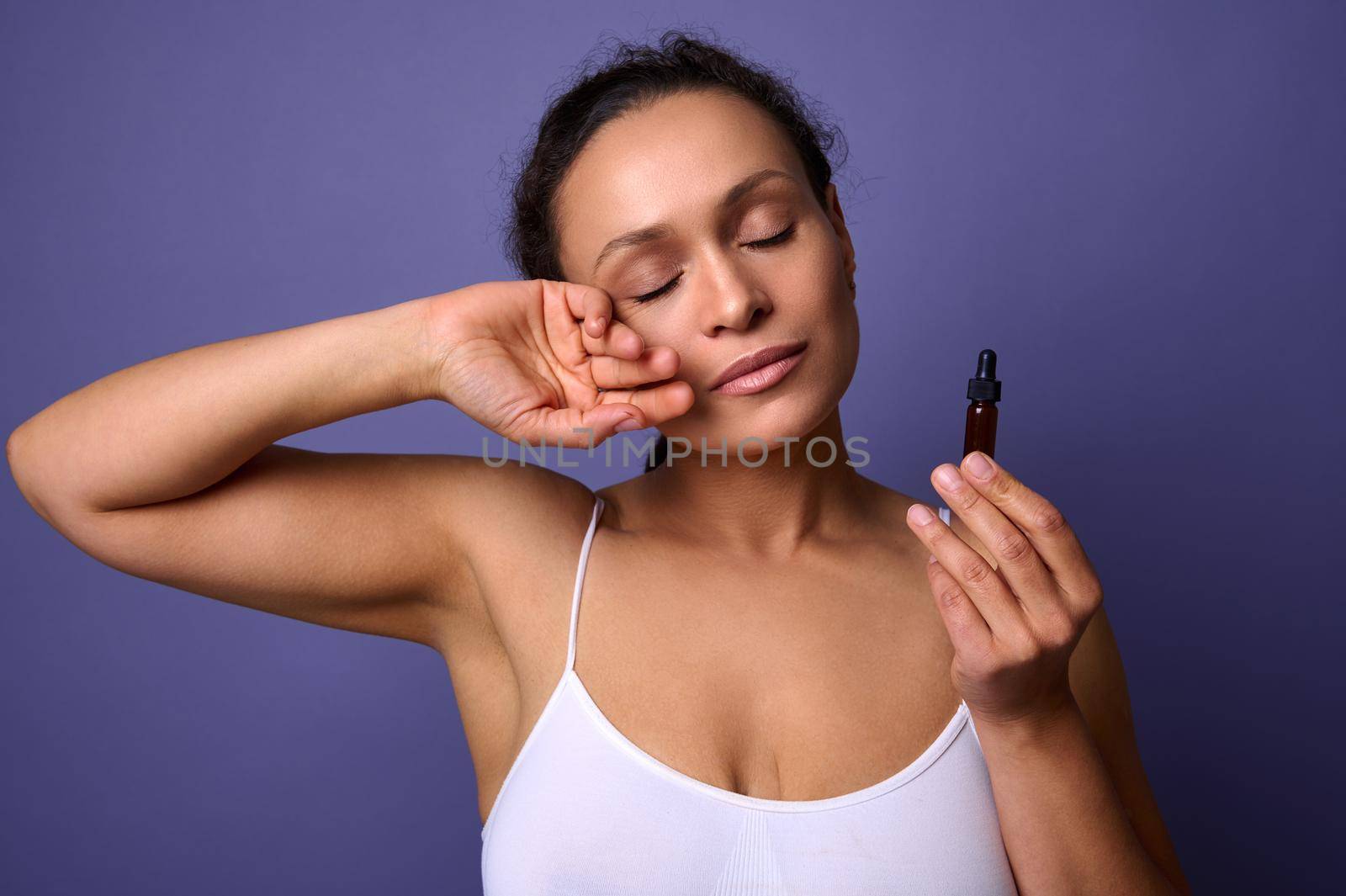 Attractive natural beauty African woman in white underwear holding jar with massage oil or smoothing anti-aging serum and posing against purple background with her eyes closed. Skin body care concept