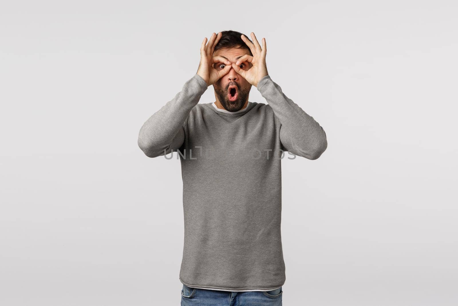 Surprised, excited and fascinated handsome bearded man in grey sweater, make circled with fingers over eyes like glasses or binocular, open mouth gasping amused, see and check out awesome promo by Benzoix