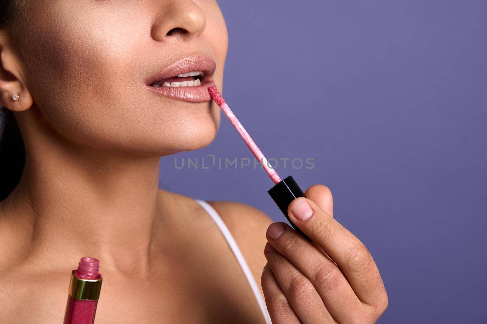 Close-up. Cropped view of attractive young woman with perfect clean skin applying lip gloss hygienic balm on plump lips isolated on purple background with copy space for advertisement. Make-up concept